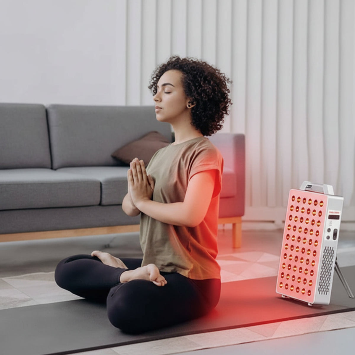 Woman meditating on a yoga mat with a pink and white device emitting light nearby in a living room.